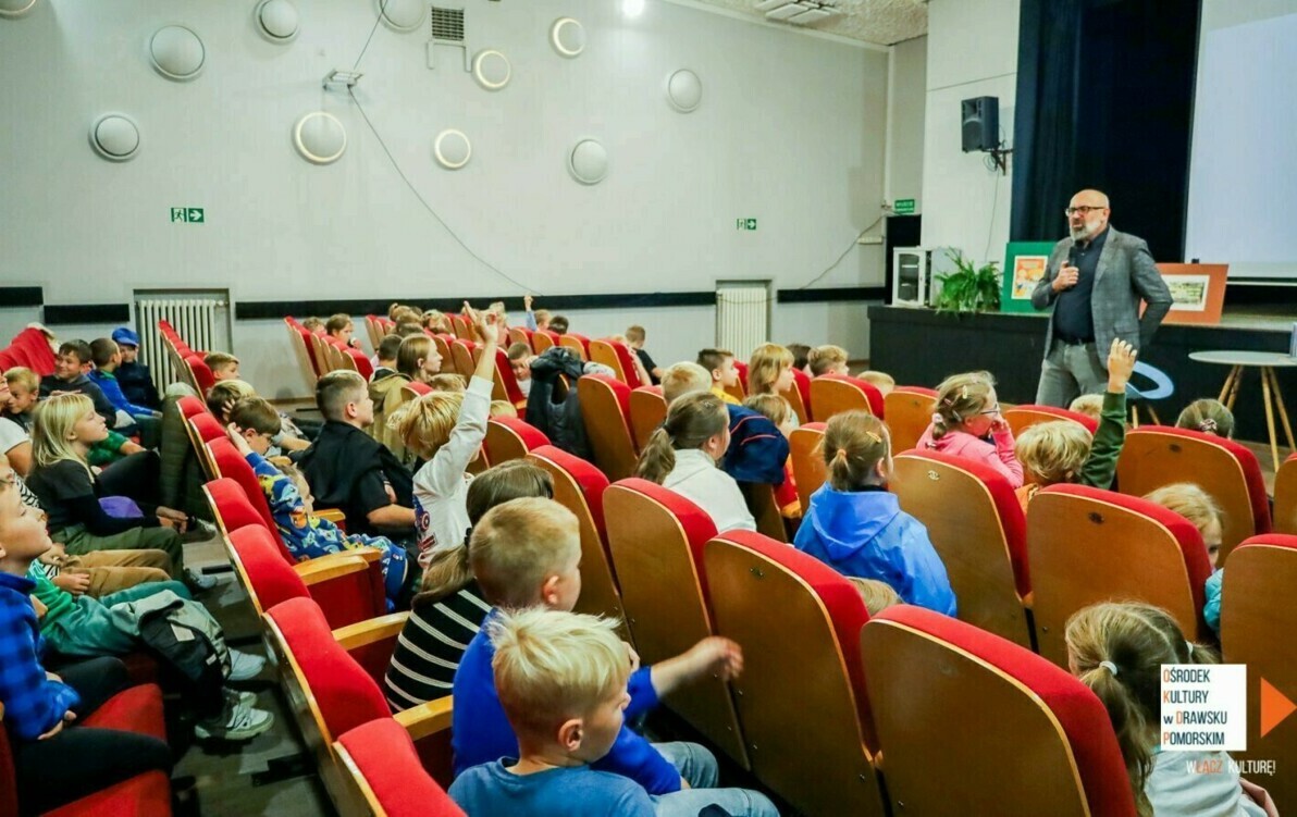 Widok na grupę dzieci siedząca przed sceną i uczestniczącą w spotkaniu.