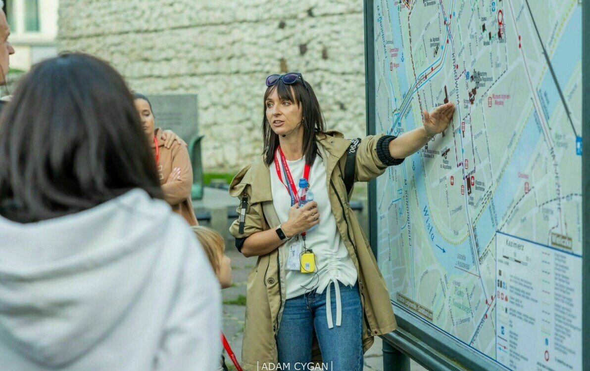 Kobieta przewodnik wskazuje ręką na mapę miasta i opowiada grupie jej słuchającej